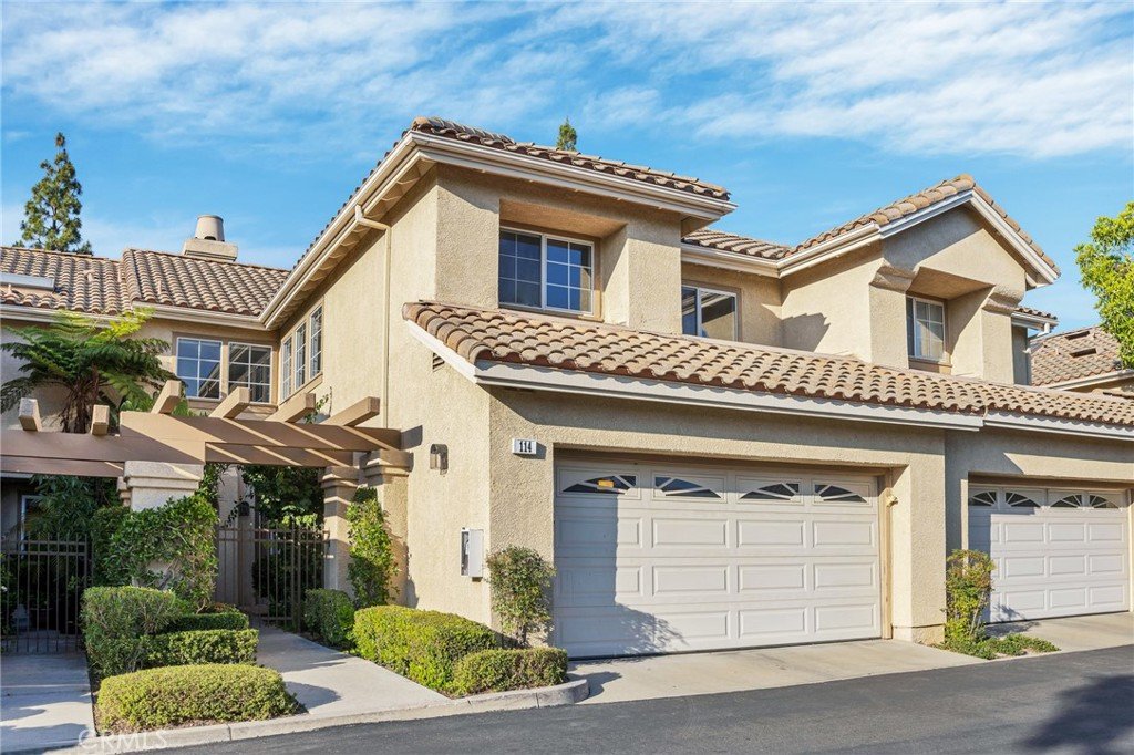 Two-story California townhouse with beige walls, tile roof, two garages, and small bushes under sunny sky.