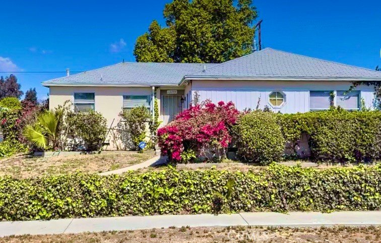 Single-story California house with light yellow walls, gray roof, greenery, and pink flowers.