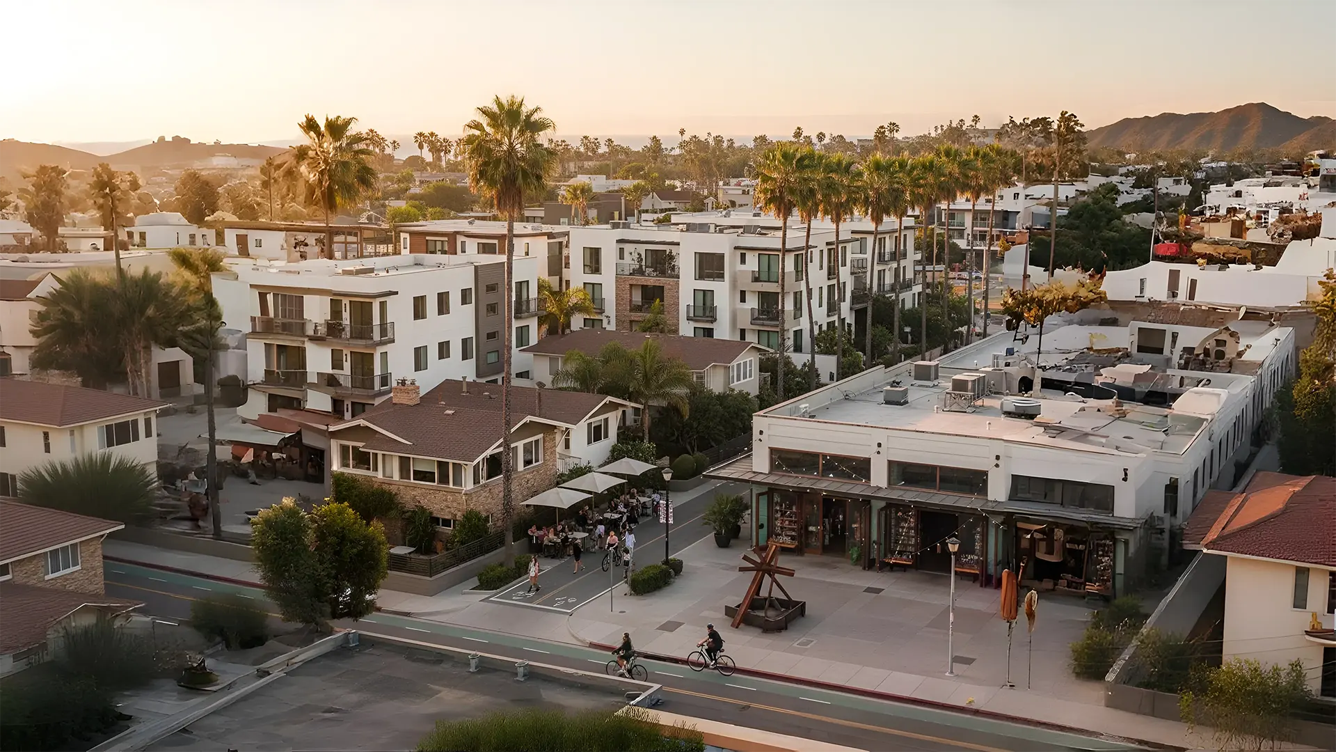 Aerial view of California neighborhood at sunset with modern apartments, palm trees, cyclists, and outdoor café.