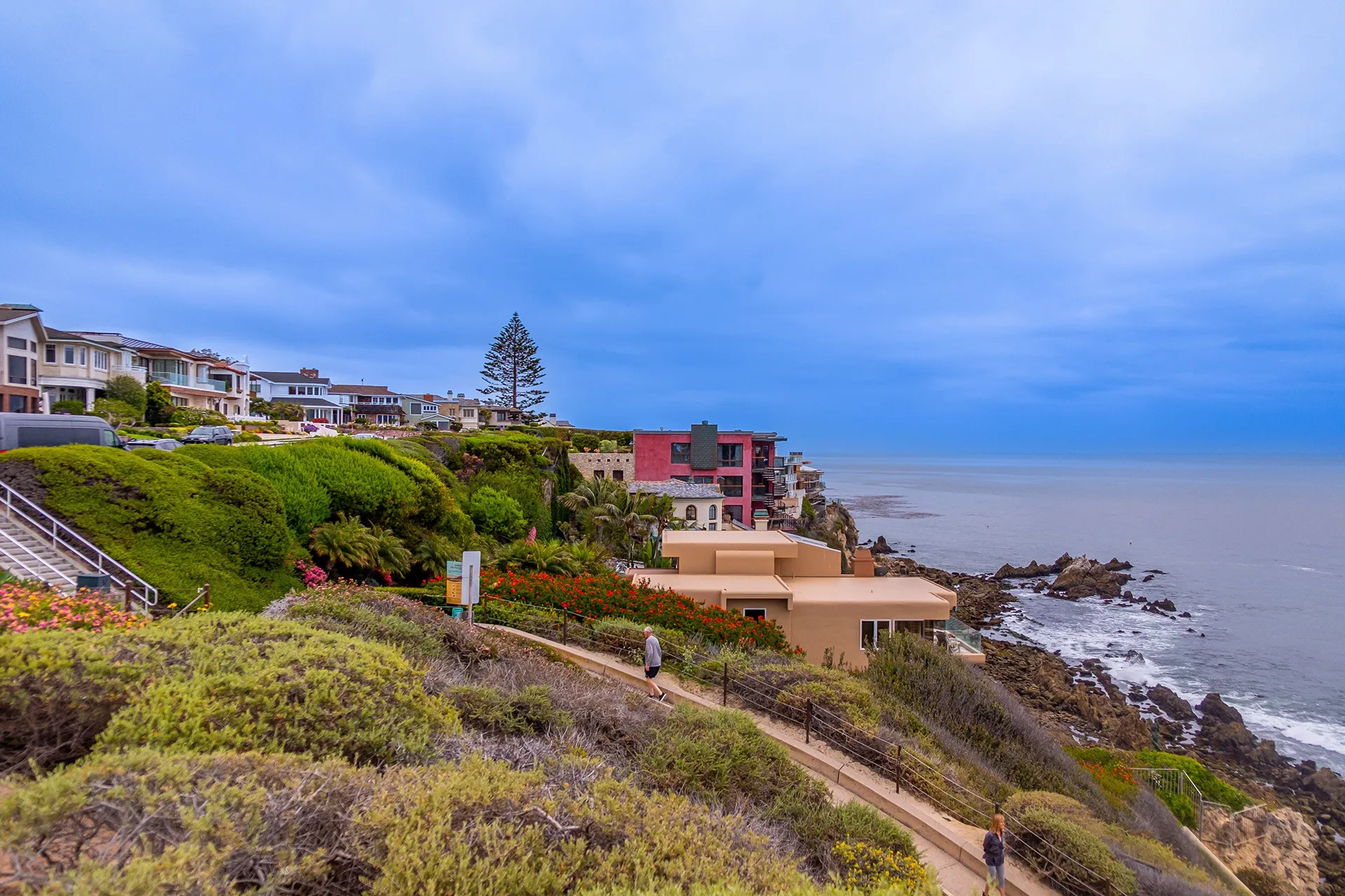 California coastal cliff view with colorful houses, greenery, winding path, and calm ocean under cloudy sky.