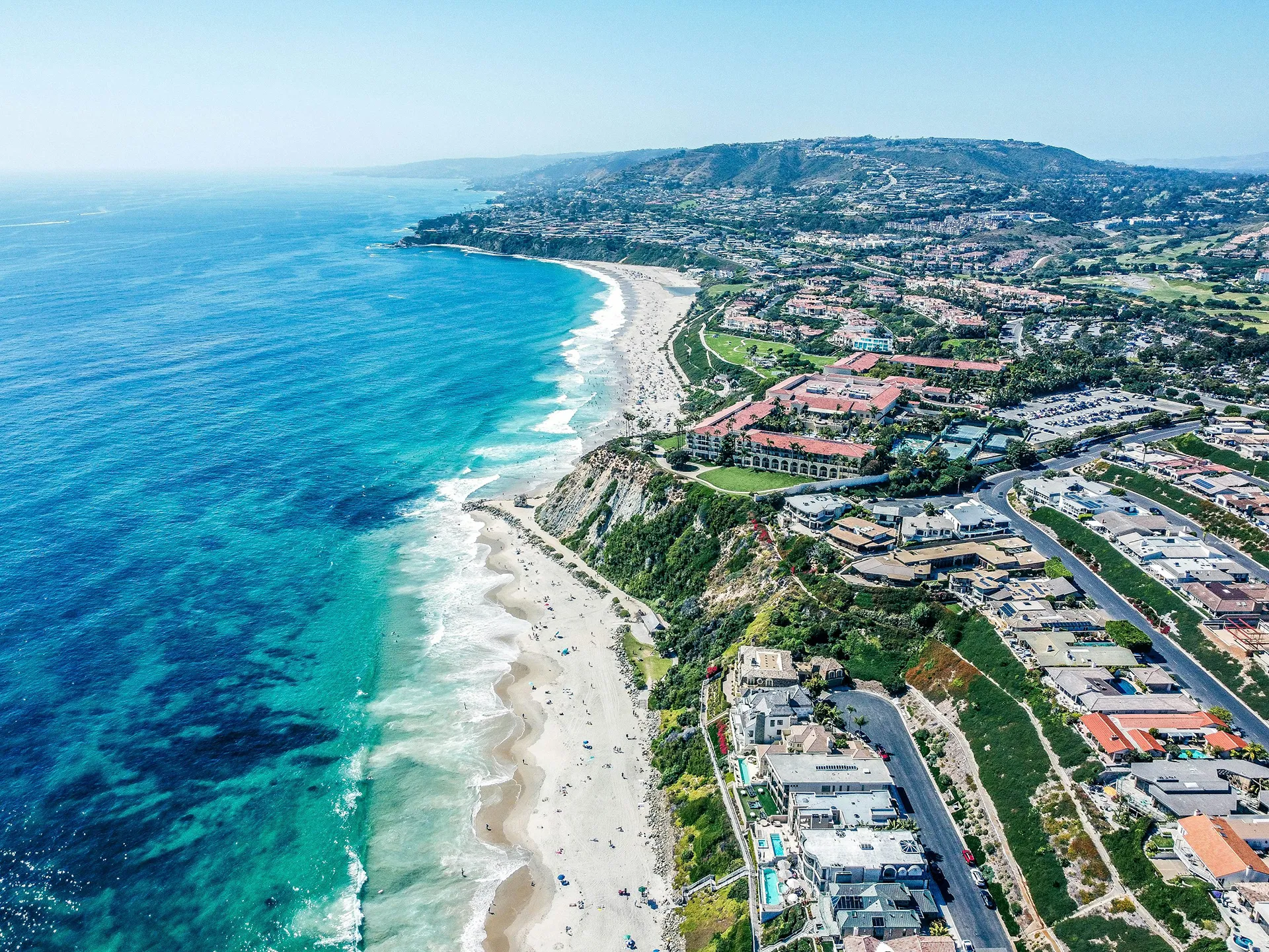 Aerial view of California coastline with sandy beach, blue waters, waves, and homes on green cliff.