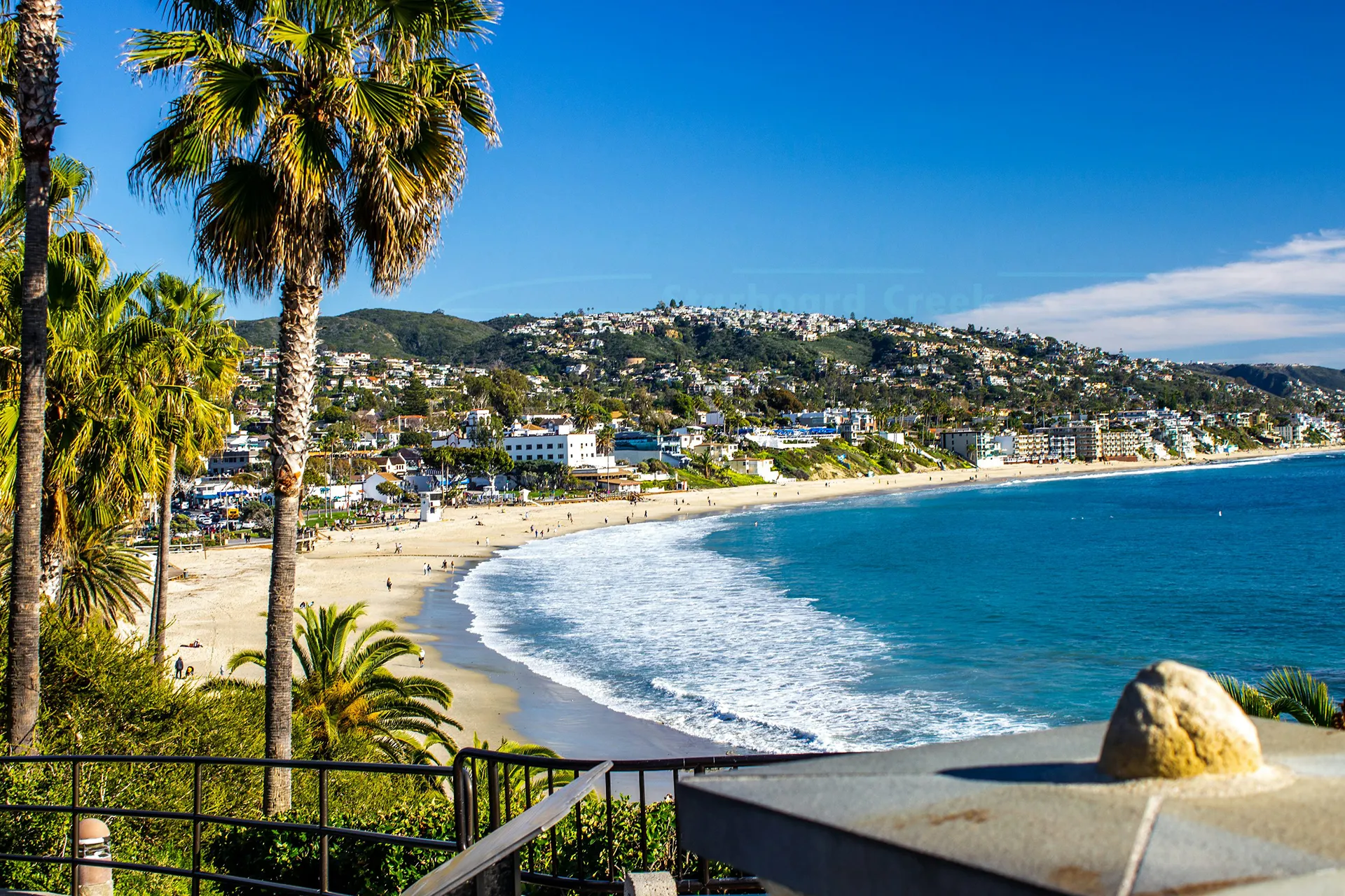 Sunny California coastal scene with palm trees, sandy beach, gentle waves, and hillside homes.